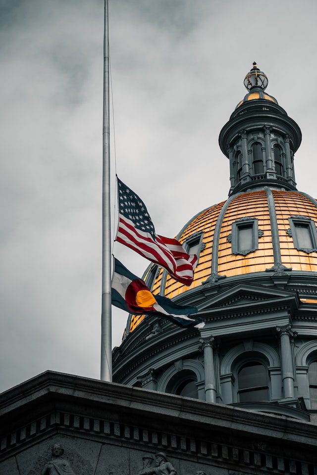The U.S. and Colorado state flags are flying at half-mast in front of the gold-domed Colorado State Capitol building under a cloudy sky.