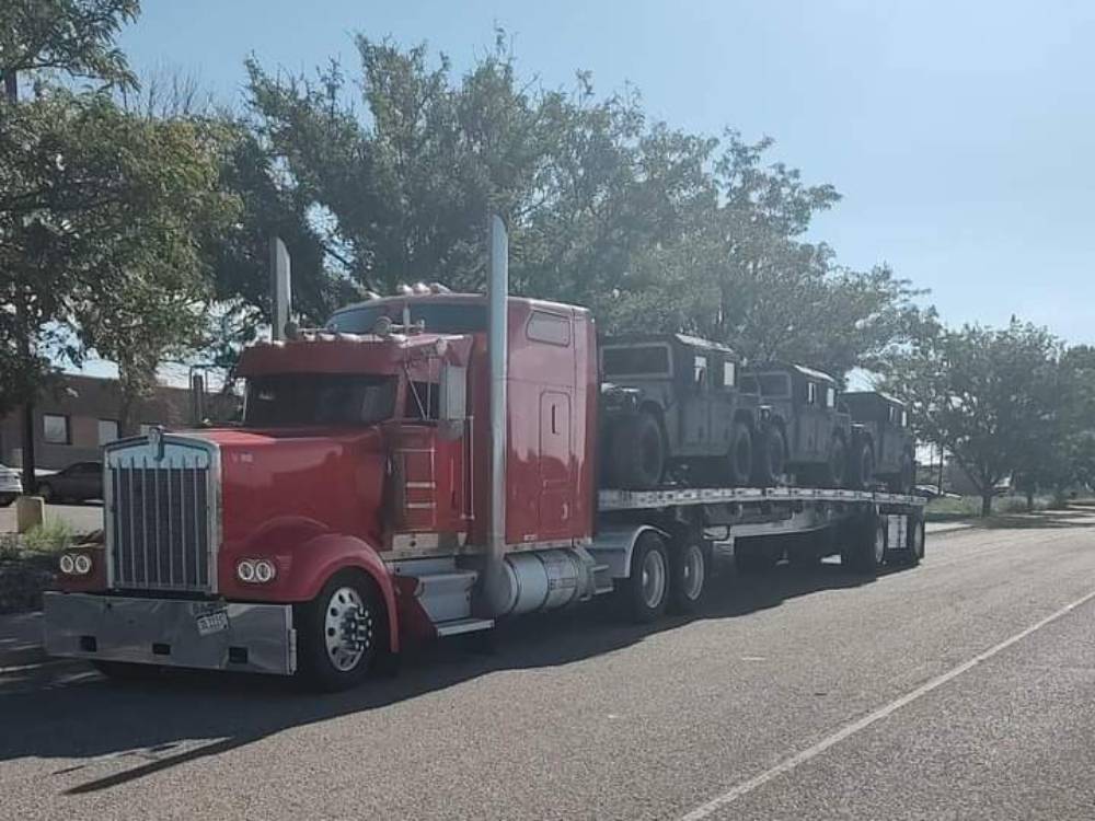 A red semi-truck is transporting three military vehicles on a flatbed trailer along a road lined with trees.