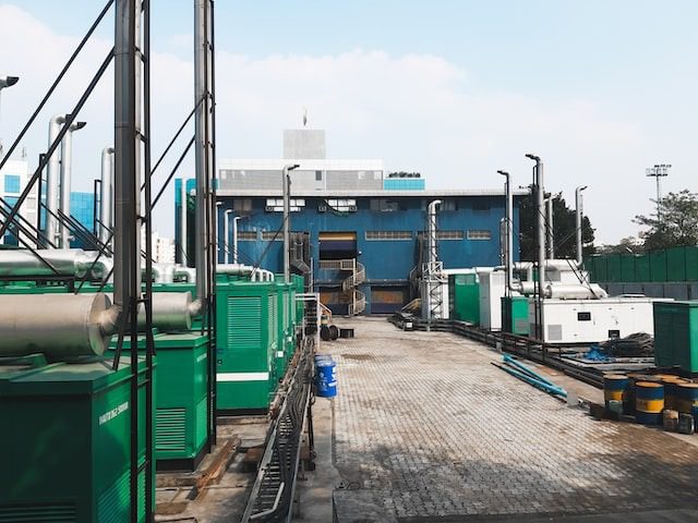 Industrial site with green machinery units, large pipes, and a blue building in the background under a clear sky.