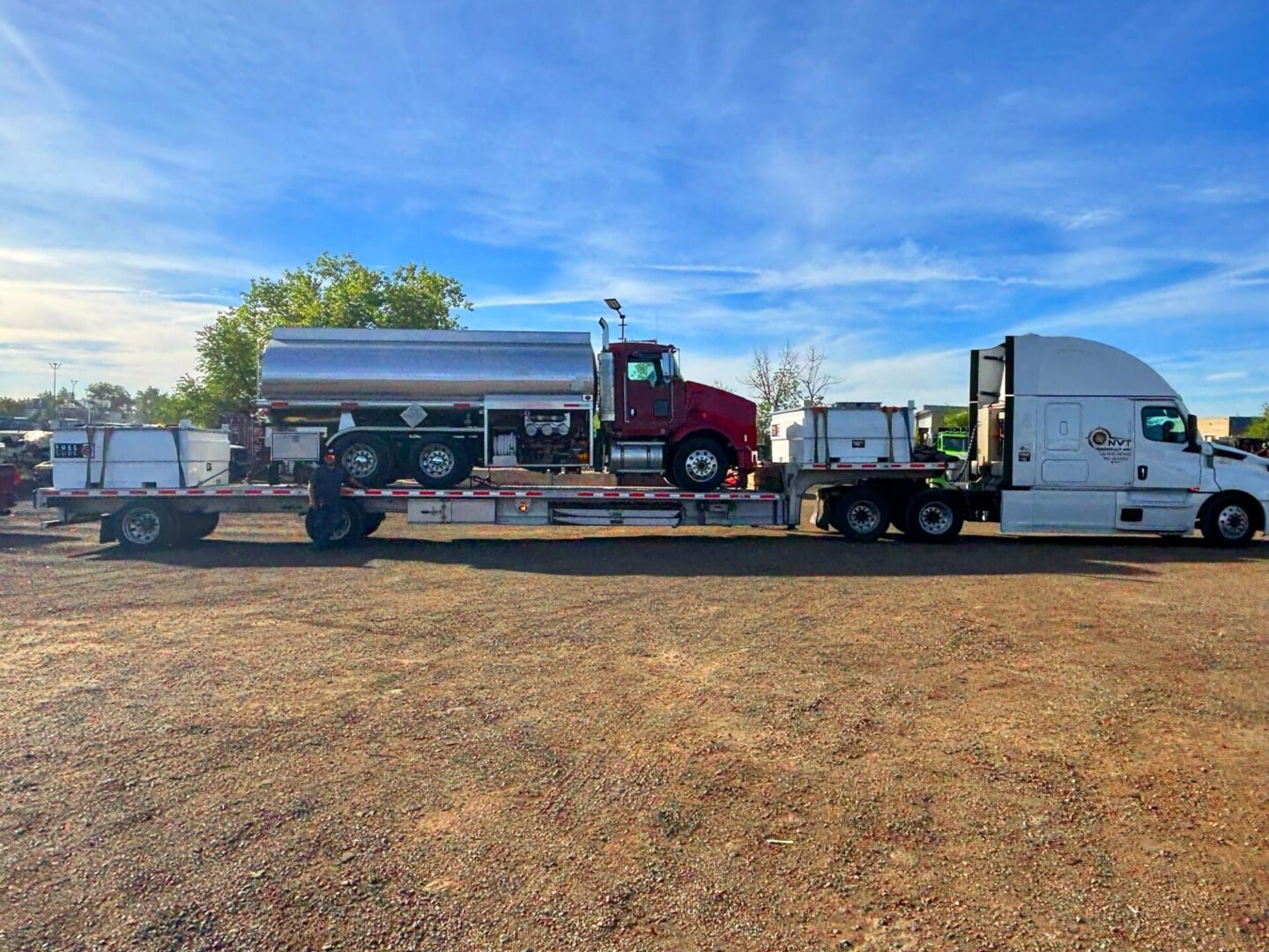 A white semi-truck and trailer carrying a red tanker truck and several white containers, parked on a gravel lot under a blue sky.
