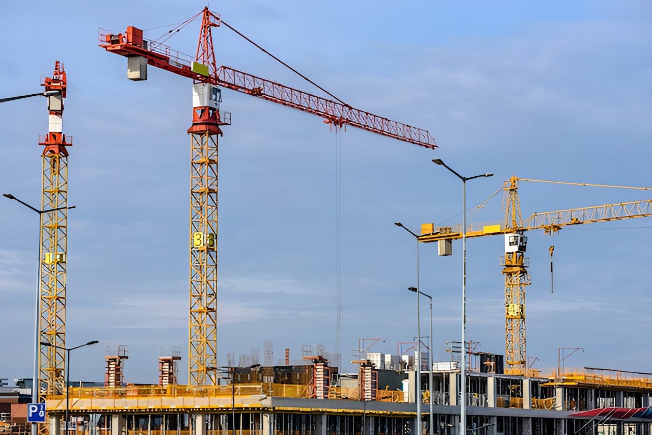 Several tall construction cranes operate above a partially completed building at a construction site under a clear sky.
