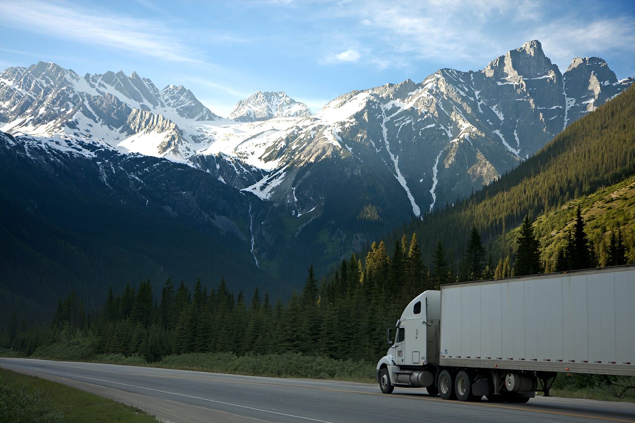 A white semi-truck drives on a road surrounded by pine trees, with snow-capped mountains and a clear sky in the background.