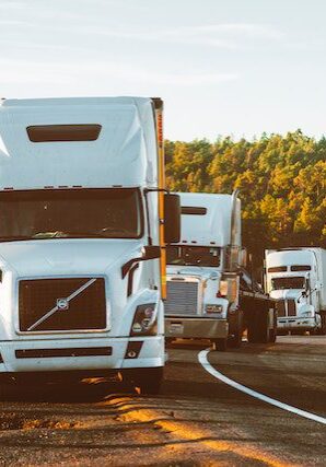 Three semi-trucks drive along a curving highway bordered by trees and shrubs, with sunlight illuminating the scene.