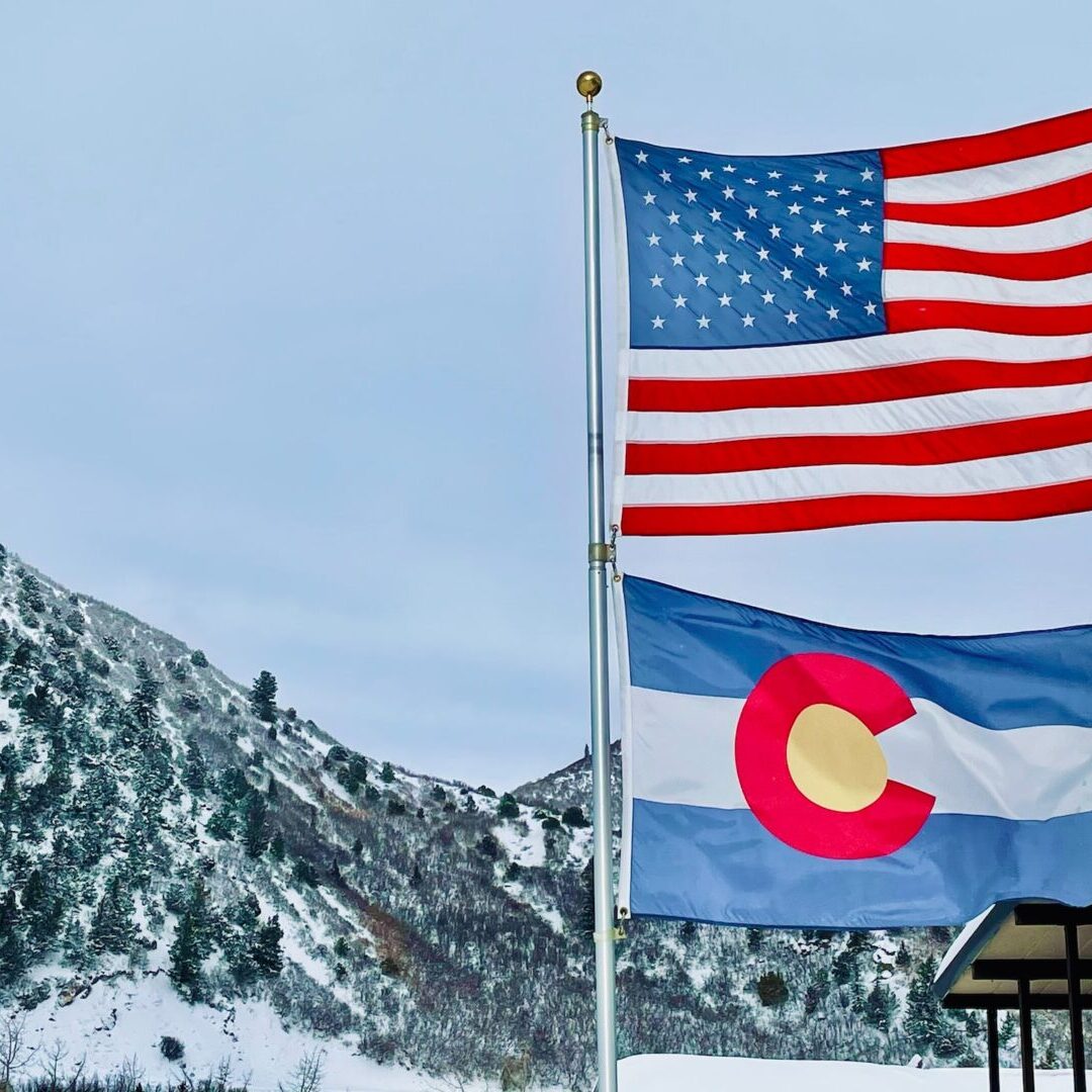 Two flags, the United States flag and the Colorado state flag, fly on a flagpole in front of a snow-covered mountain landscape.