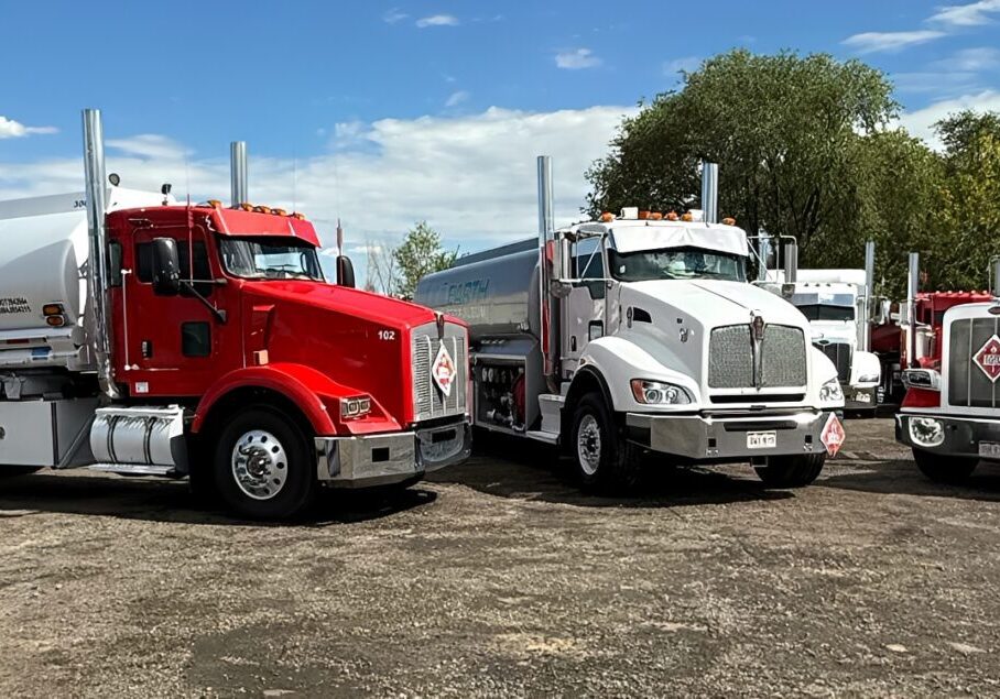 Three large fuel tanker trucks are parked side by side in a lot on a sunny day, with trees and other trucks visible in the background.